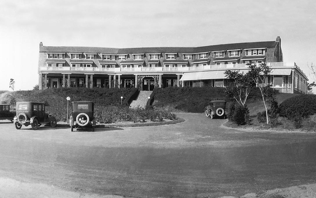 A large historic building with multiple windows and vintage cars parked in front, captured in black and white.