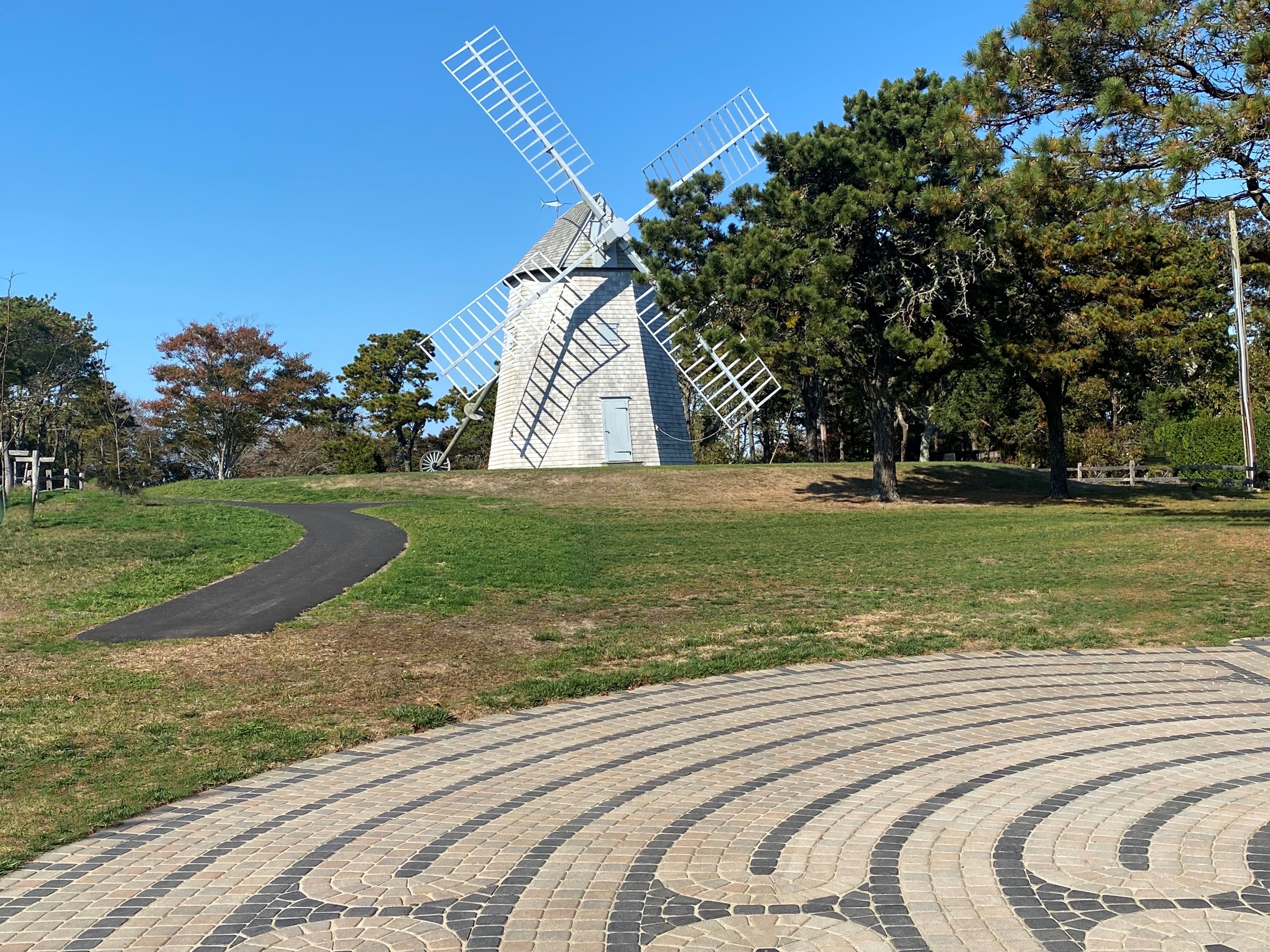 A scenic view of a white windmill surrounded by greenery and trees, with a paved path and a patterned circular area in the foreground.
