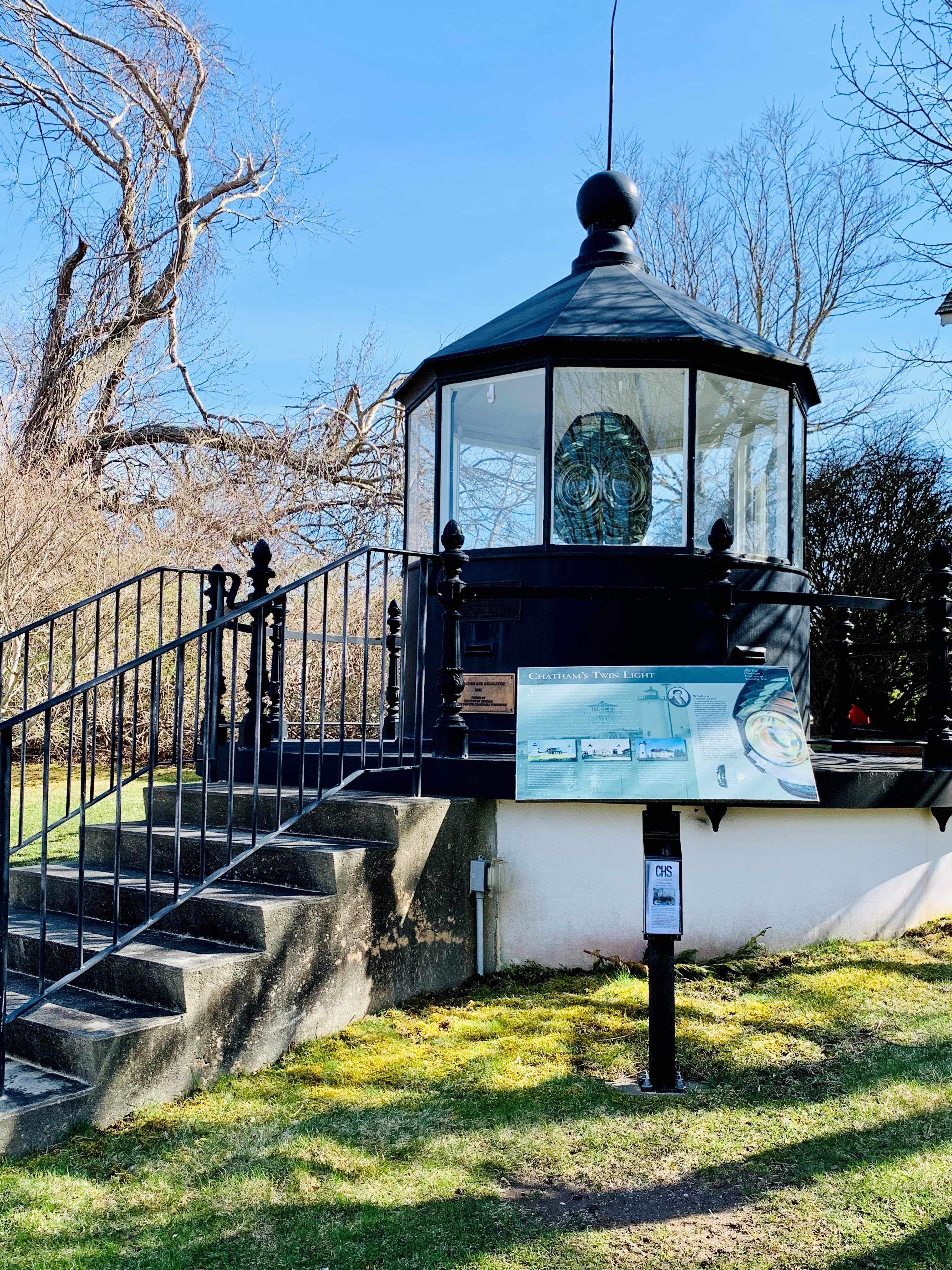 A historic lantern room on a platform, surrounded by railings with an information sign about Chatham's Twin Light.