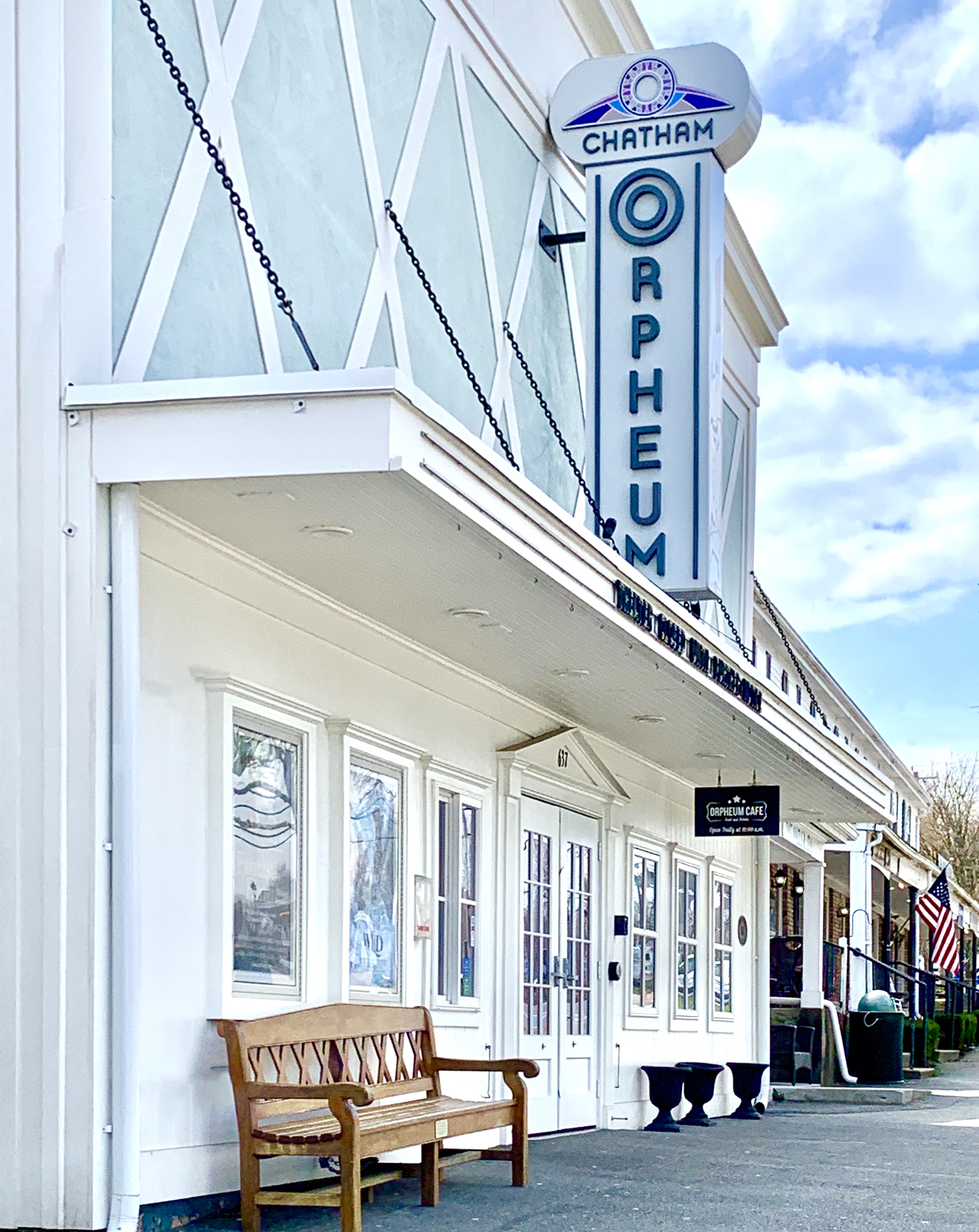 A building with a sign for "Chatham Orpheum," featuring a bench outside and a flag in the background, under a partly cloudy sky.