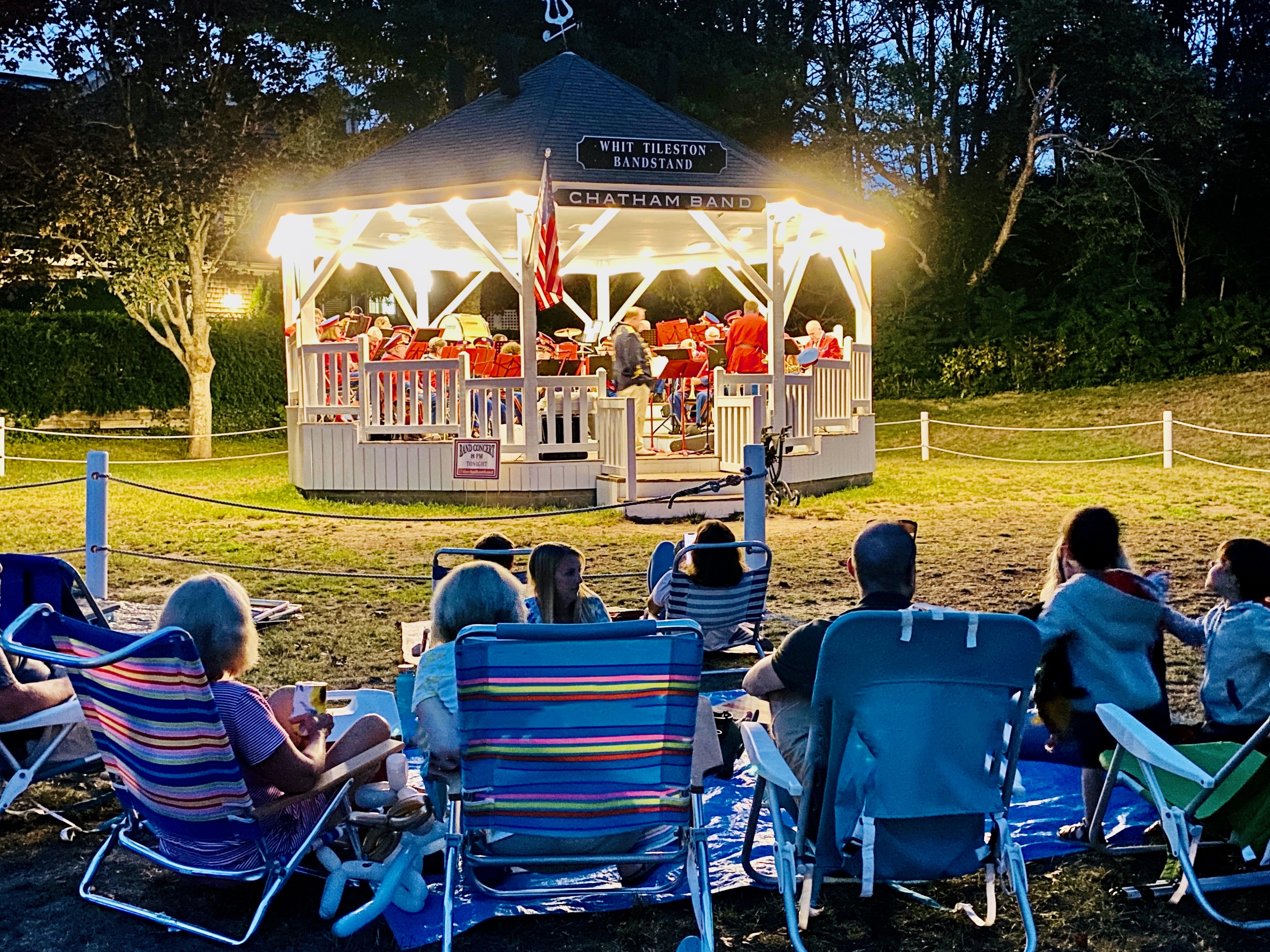 A family enjoys a live band performance at an illuminated bandstand in a park, seated in colorful lawn chairs on a summer evening.
