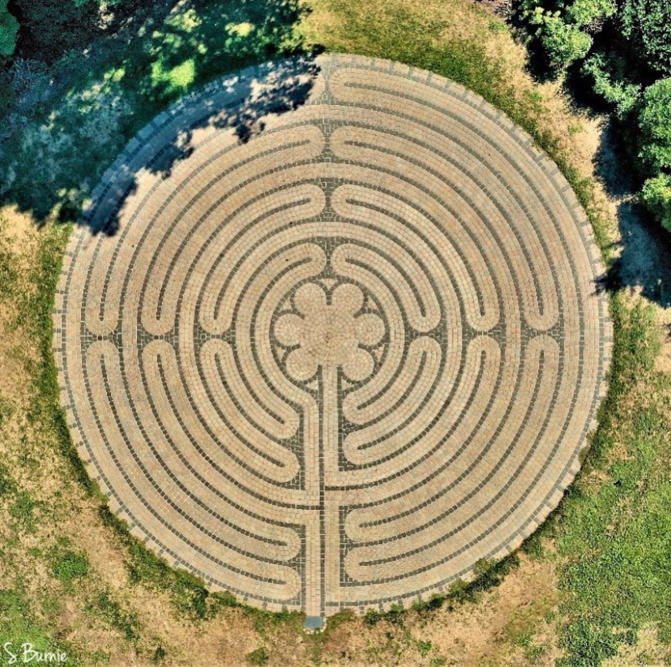The image shows a circular labyrinth made of stone pathways set in a grassy area, bordered by trees on one side.