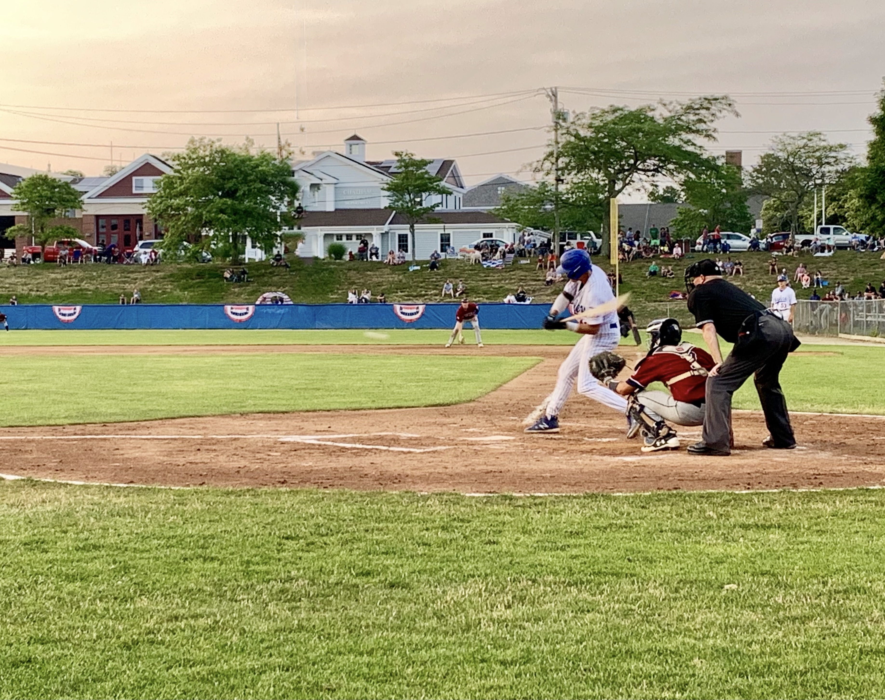 A baseball game in progress, with a batter, catcher, and umpire at home plate, spectators watching from the background.