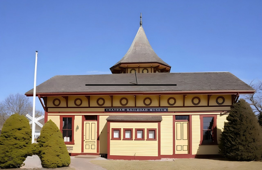A historic building with a pointed roof, labeled "Railroad Museum," surrounded by bushes and a pathway under a clear blue sky.