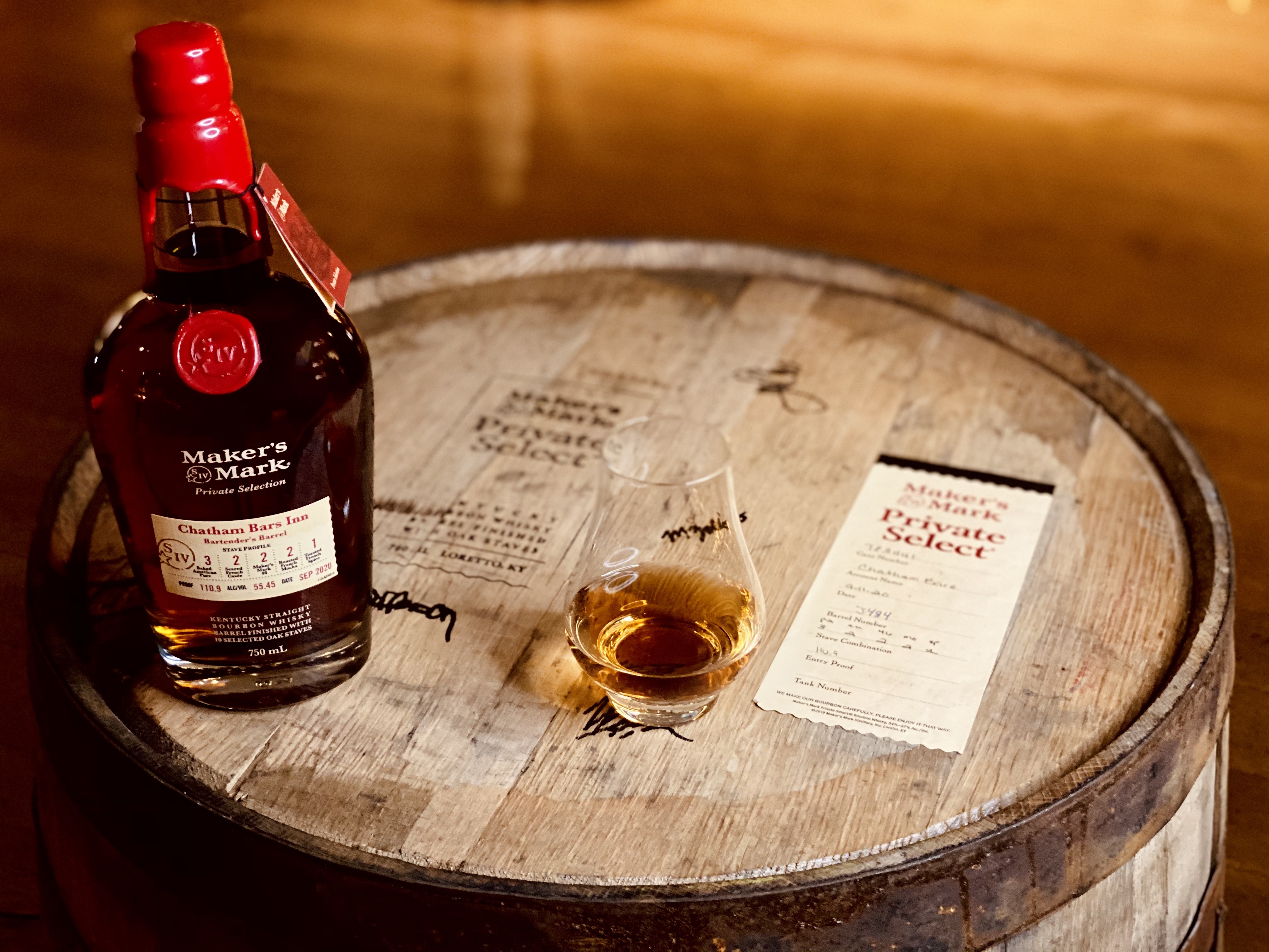 A Maker's Mark Private Selection bottle on a wooden barrel with a tasting glass and a menu or label beside it.