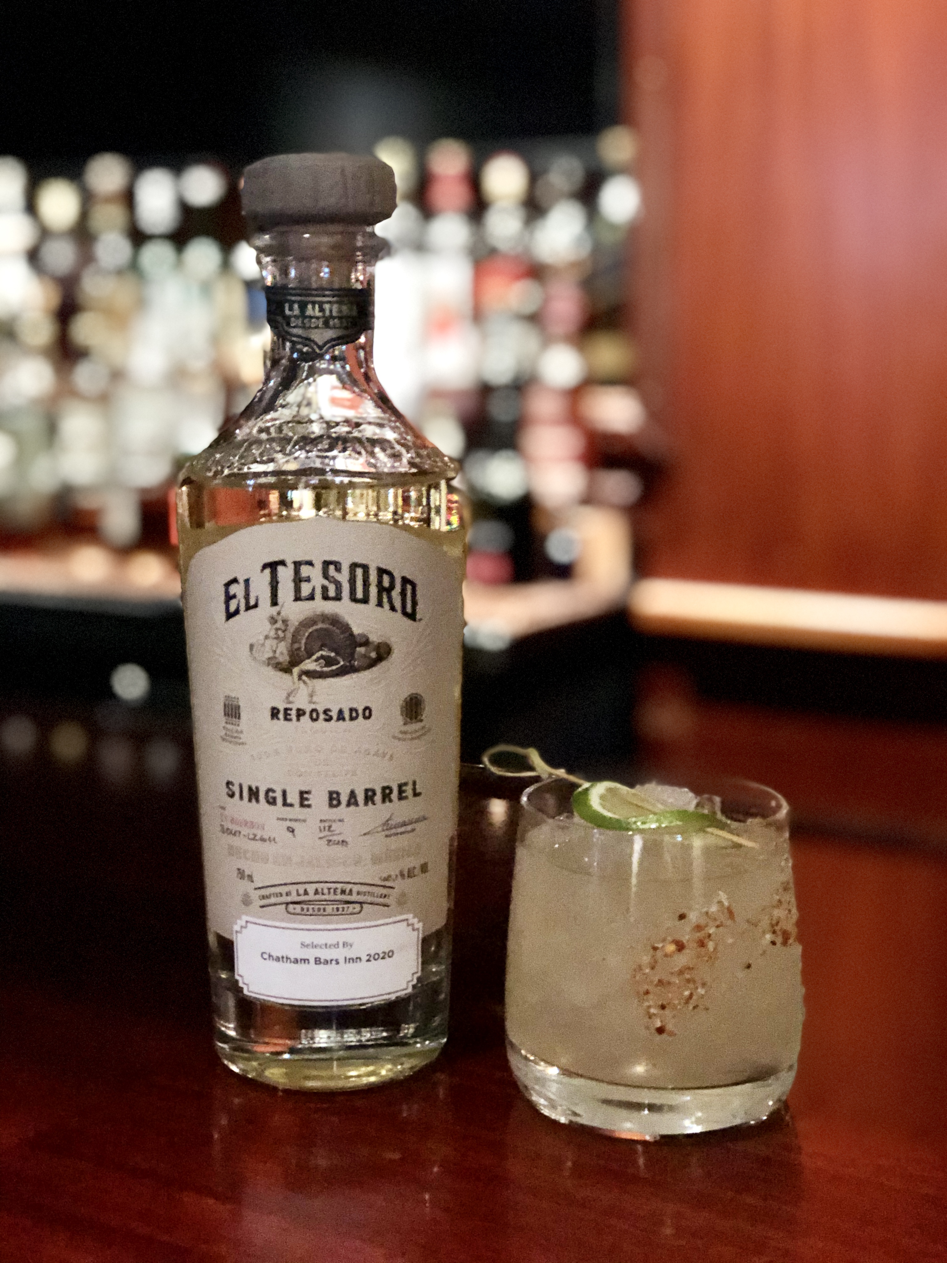 A bottle of El Tesoro Reposado tequila and a cocktail with lime and a spicy rim on a bar counter.