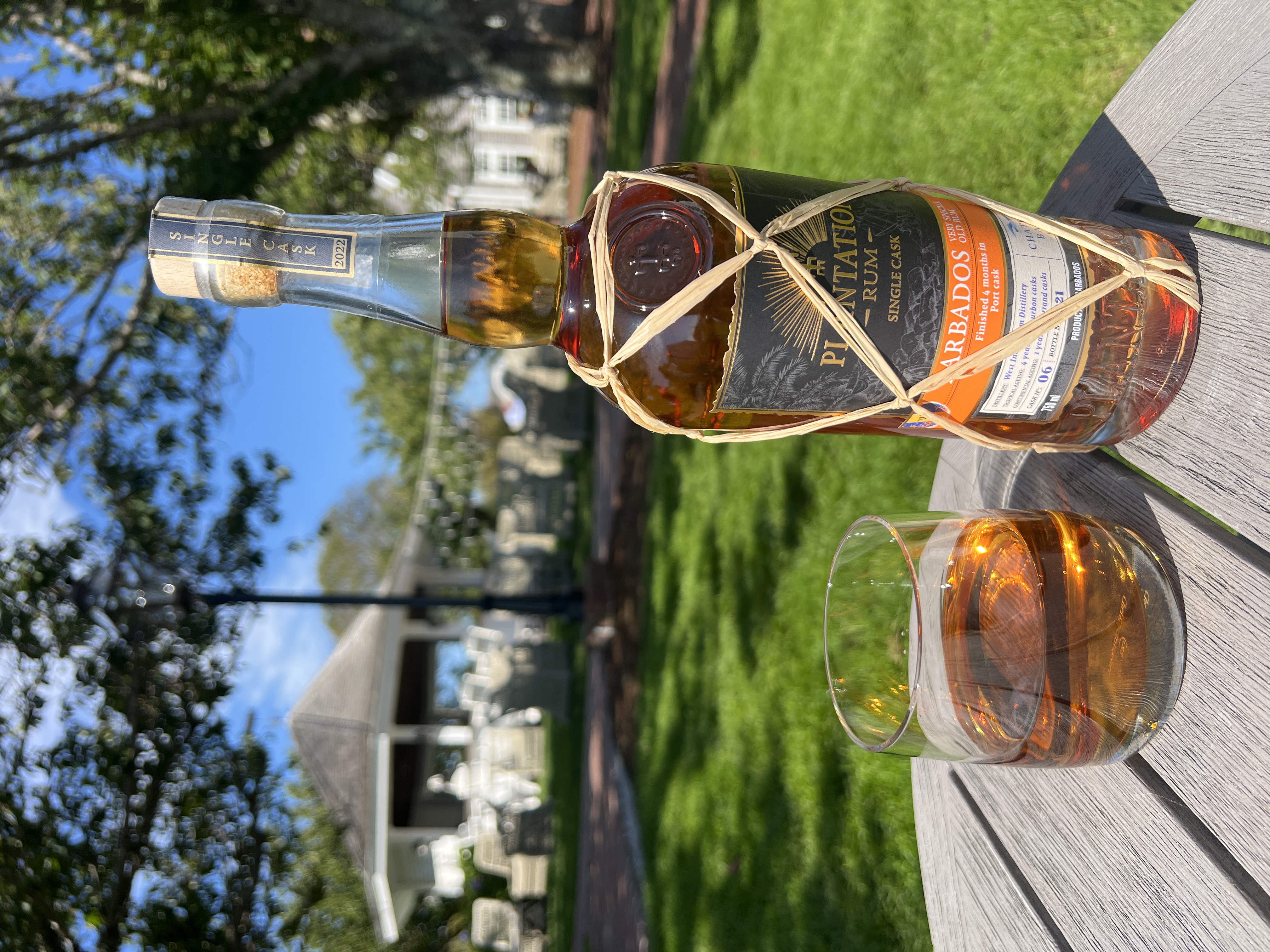A bottle and glass of Barbados Plantation Rum on a wooden table outdoors, with a gazebo in the background.