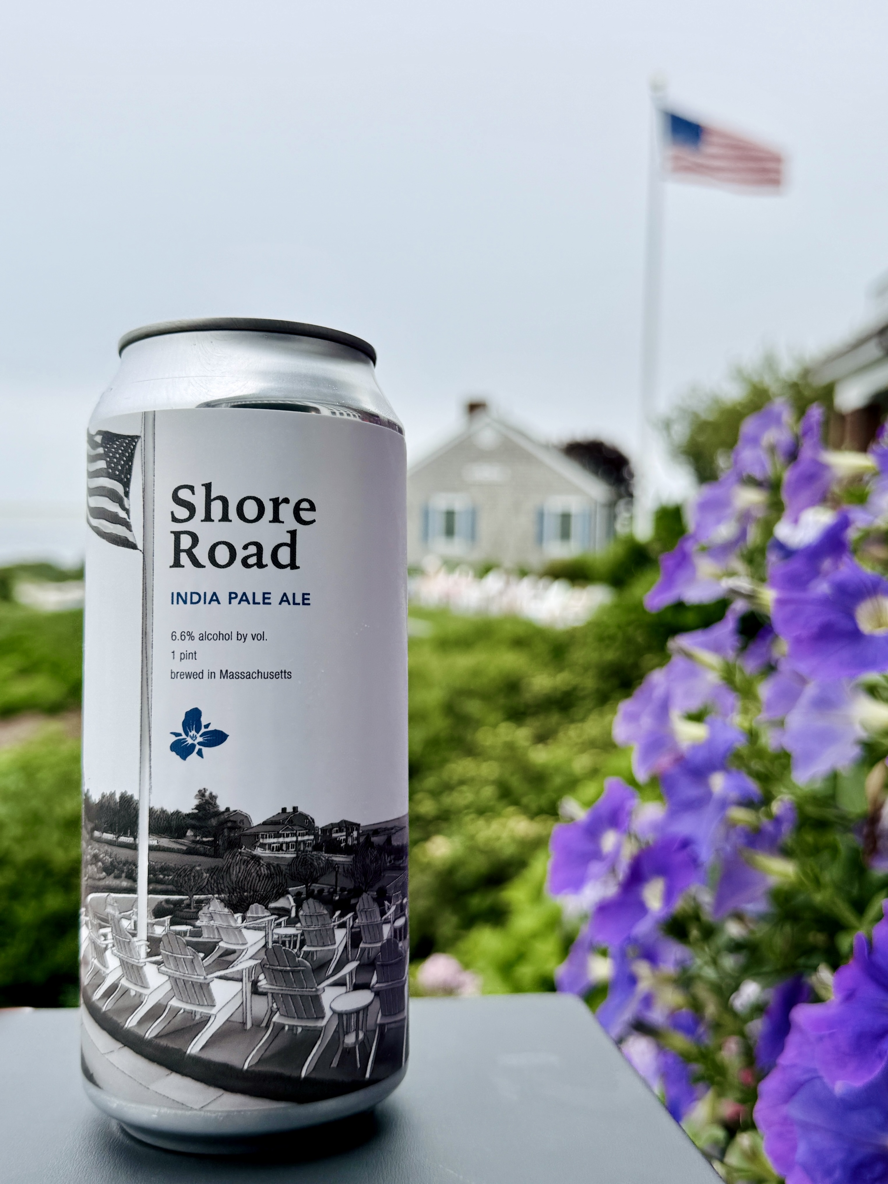 A can of Shore Road India Pale Ale is in the foreground, with purple flowers and an American flag flying in the background.