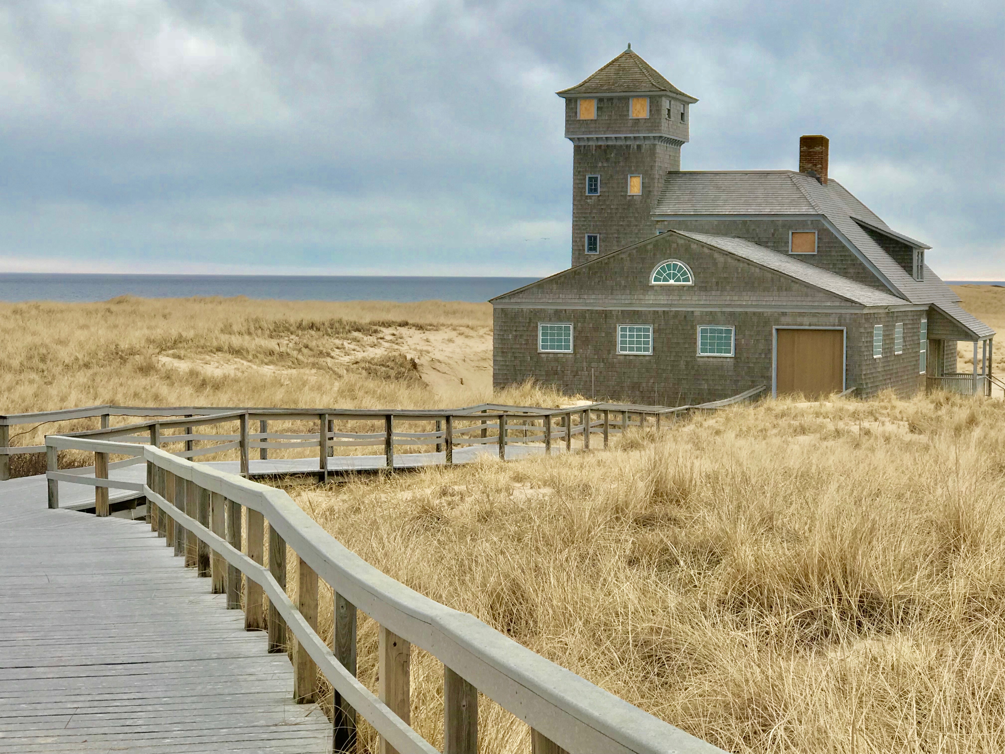 A wooden walkway leads through dunes to a brown building with a tower, under a cloudy sky by the sea.