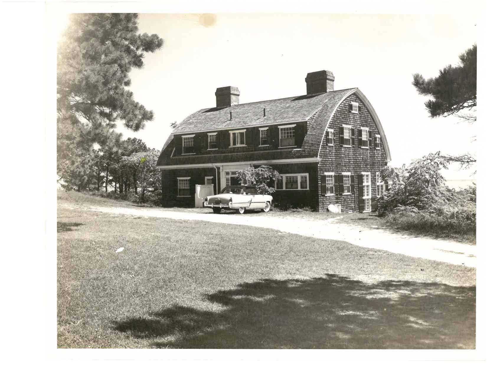 A classic car is parked in front of a rustic, shingled house surrounded by trees, with a lawn and rural setting.