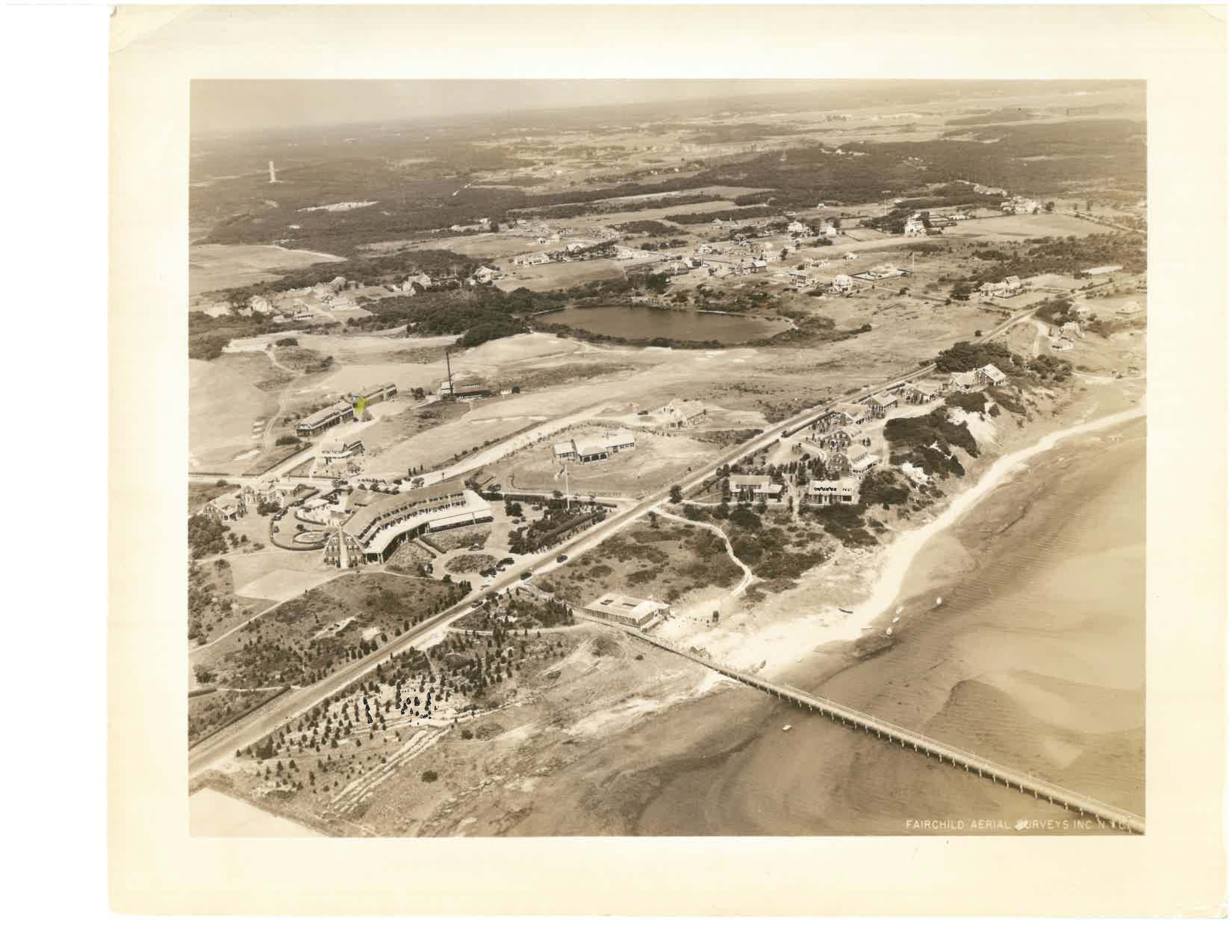 Aerial view of a coastal area with buildings, roads, a pier, and a beach, surrounded by landscape and water.