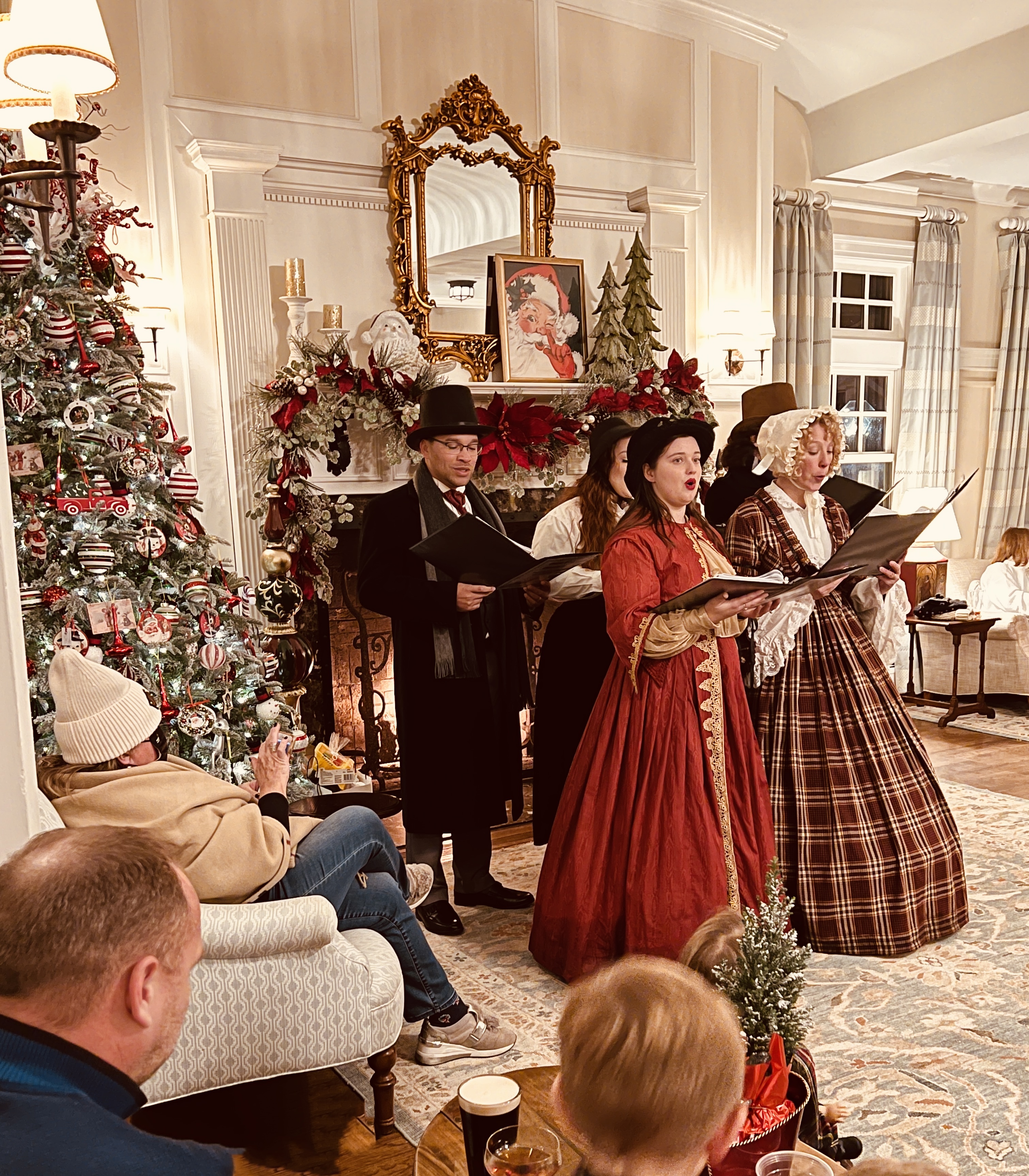 A group in period costumes sings carols by a decorated fireplace, with onlookers enjoying the festive scene, including a Christmas tree.