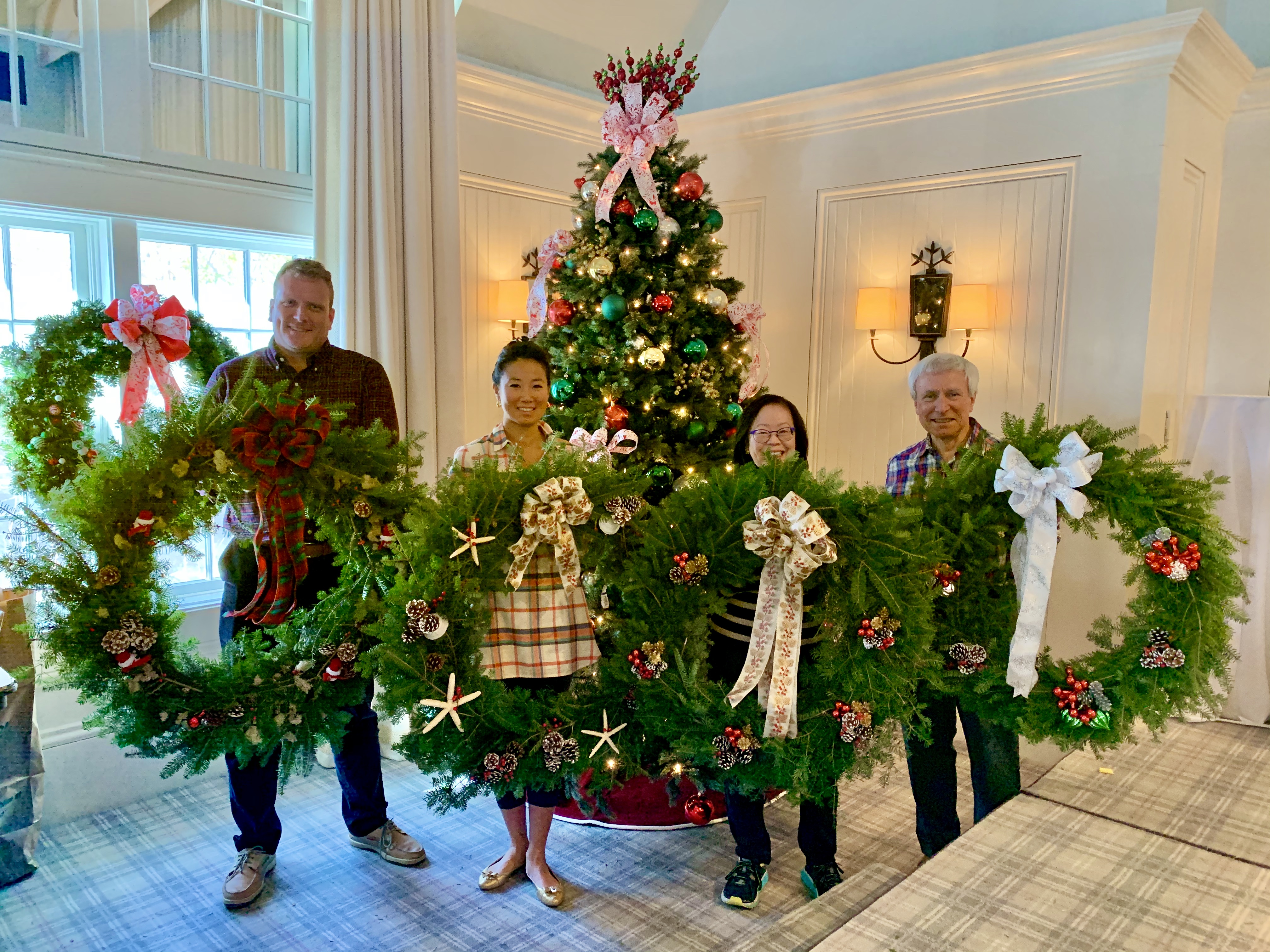 Four people hold a large Christmas wreath in front of a decorated tree indoors.