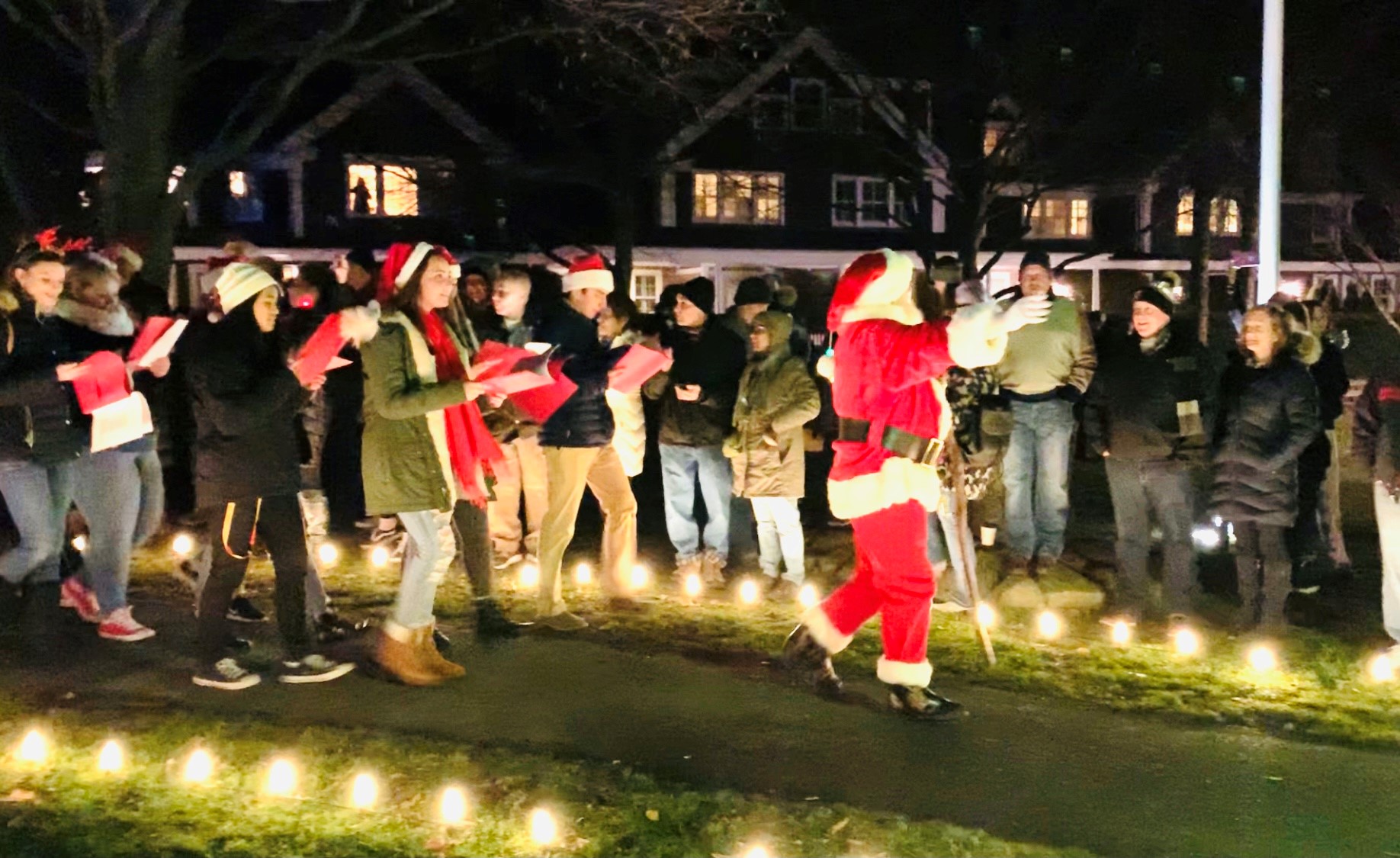 A group dressed in festive attire is singing carols outdoors at night, surrounded by lit candles and onlookers.
