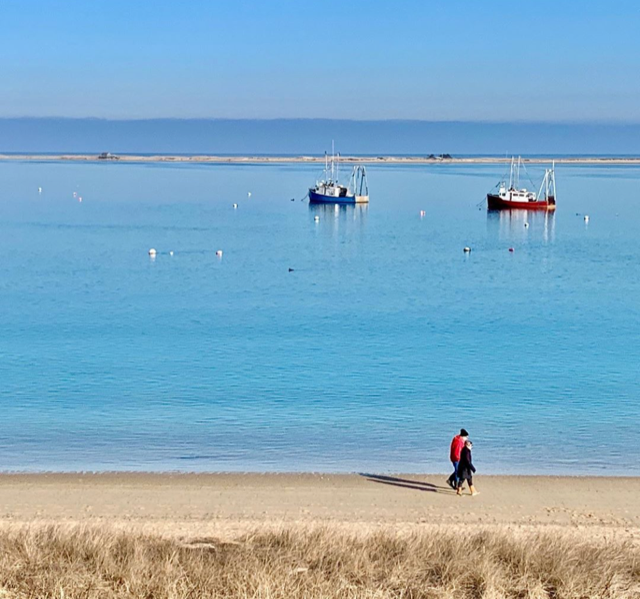 A calm beach scene with two people walking on the sand, boats anchored on clear blue water, and a serene horizon.