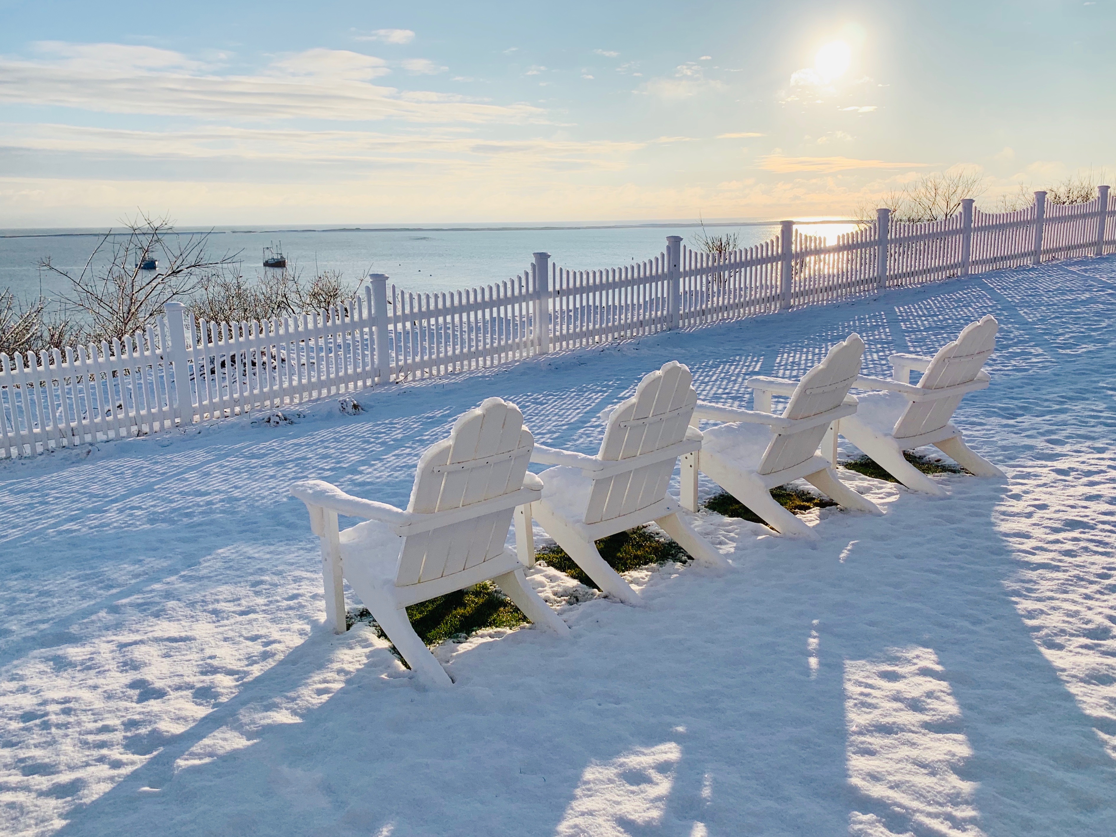 Four white Adirondack chairs on snow face a sunny, waterfront view with boats and a picket fence.