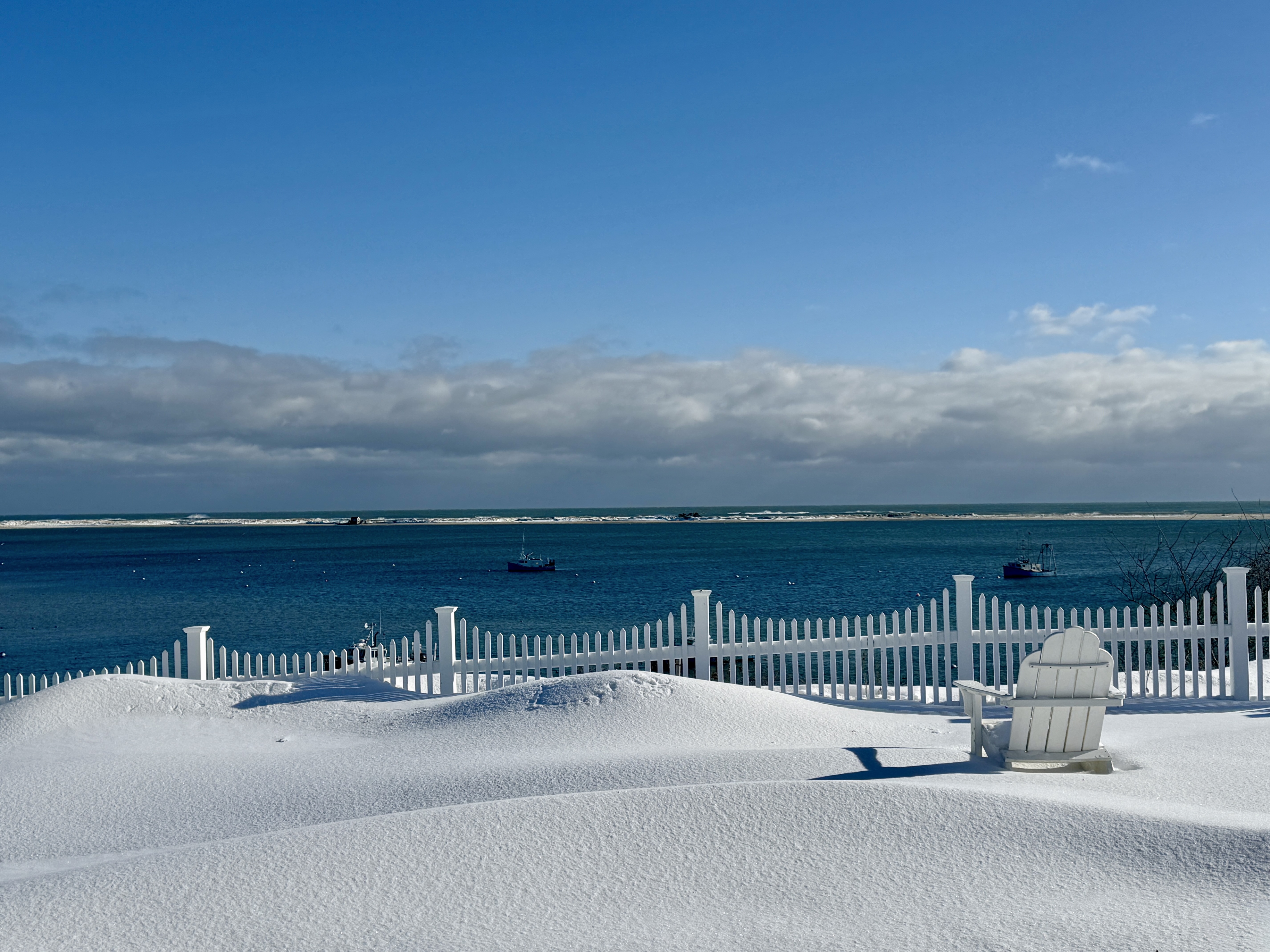 A snowy beach scene with a white picket fence, an Adirondack chair, and boats on the water under a blue sky.