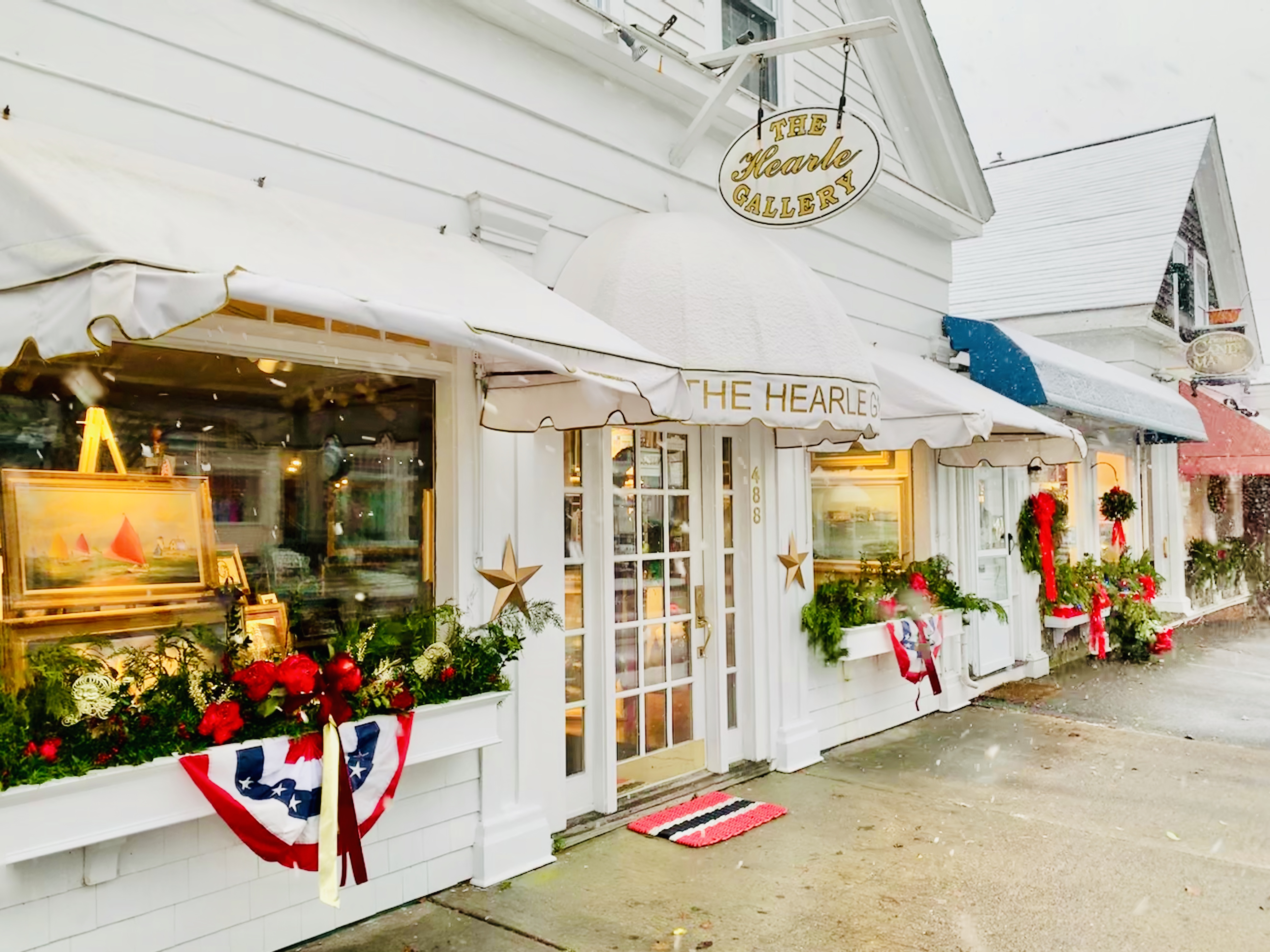 A decorated art gallery storefront with paintings, festive holiday wreaths, and a red, white, and blue banner outside.