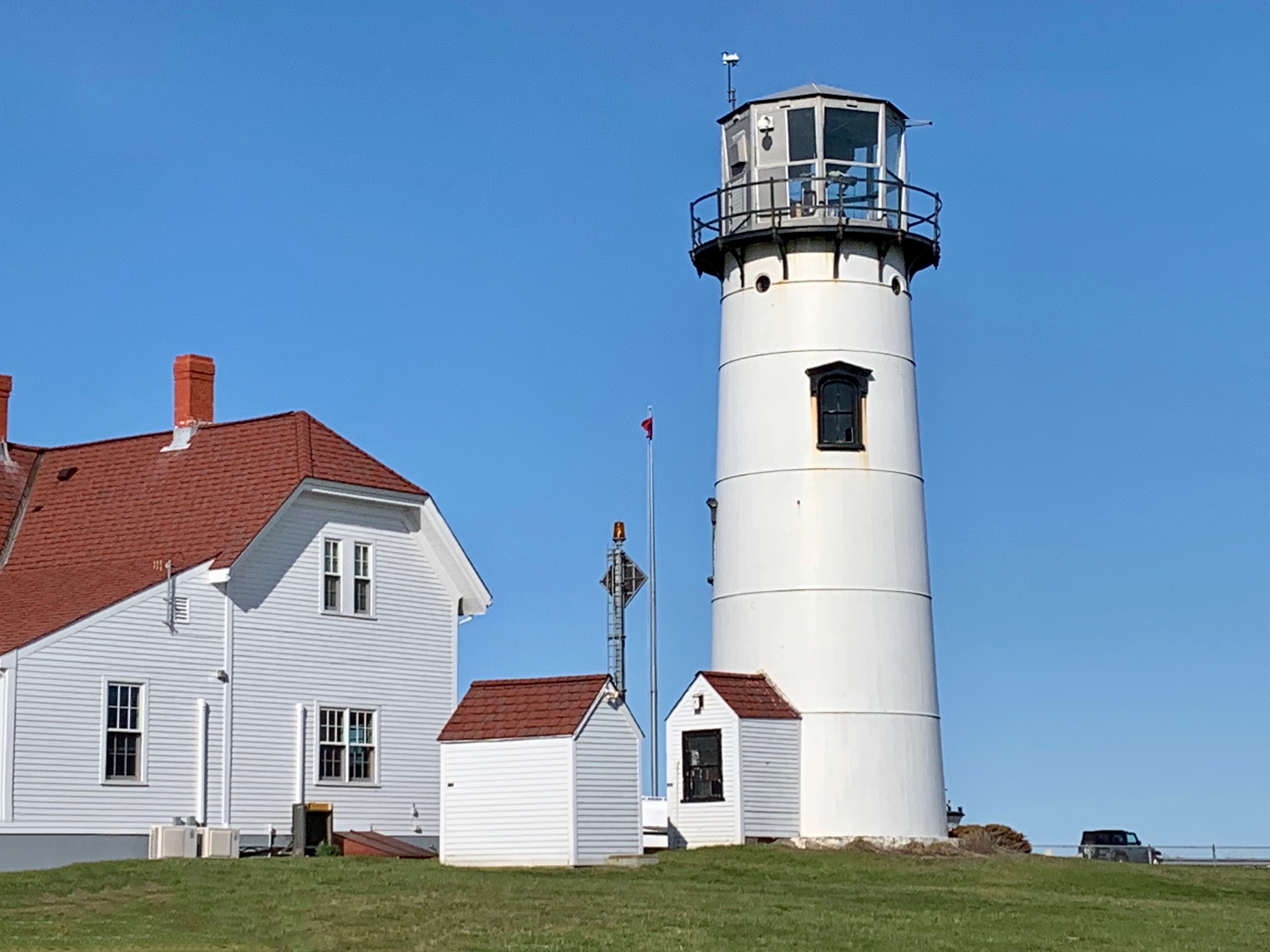 A white lighthouse with a red-roofed building, set against a clear blue sky, stands on a grassy area.