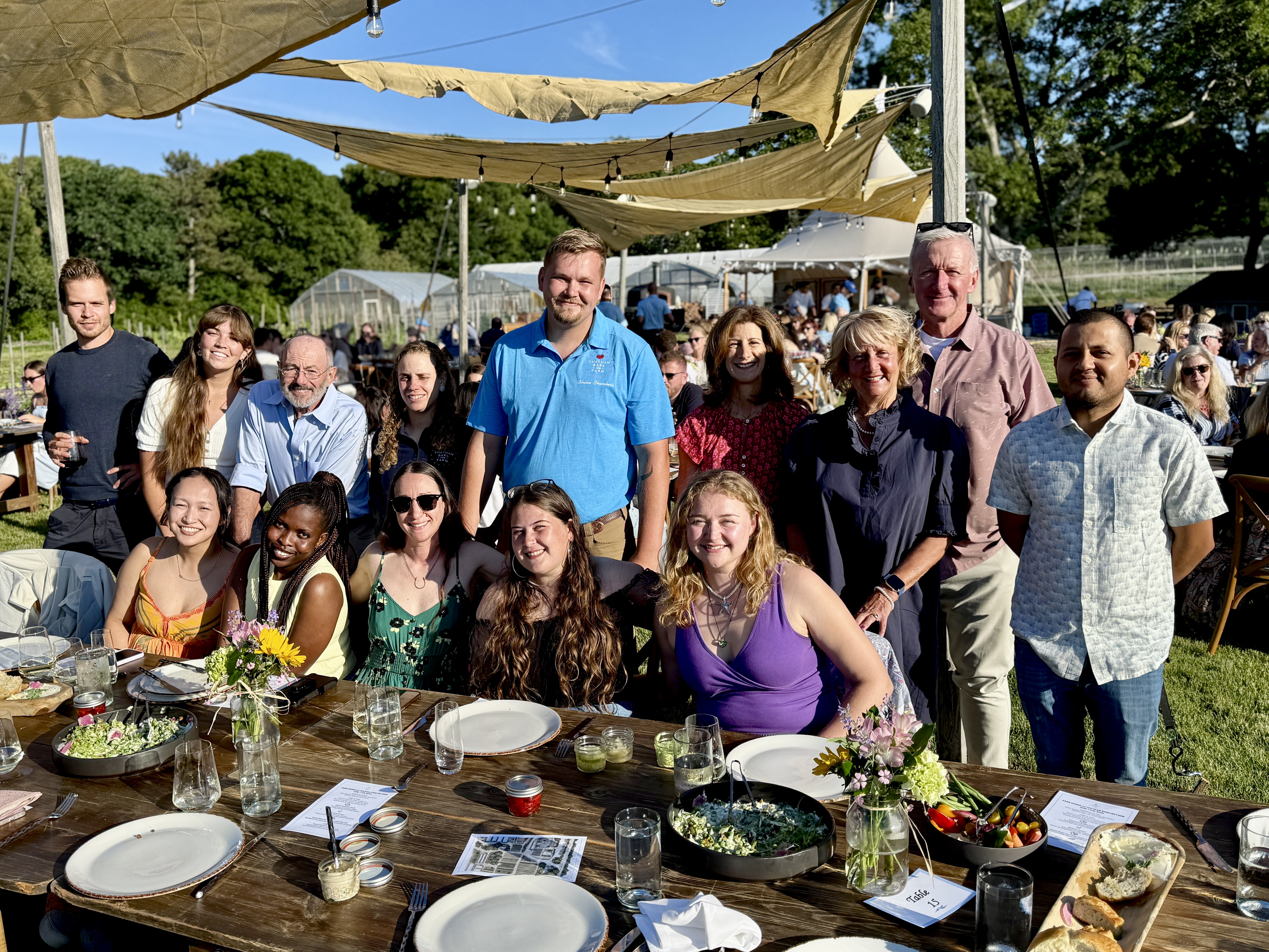 A group of people gathered outdoors around a dining table with food, under sunny skies and canvas canopies.