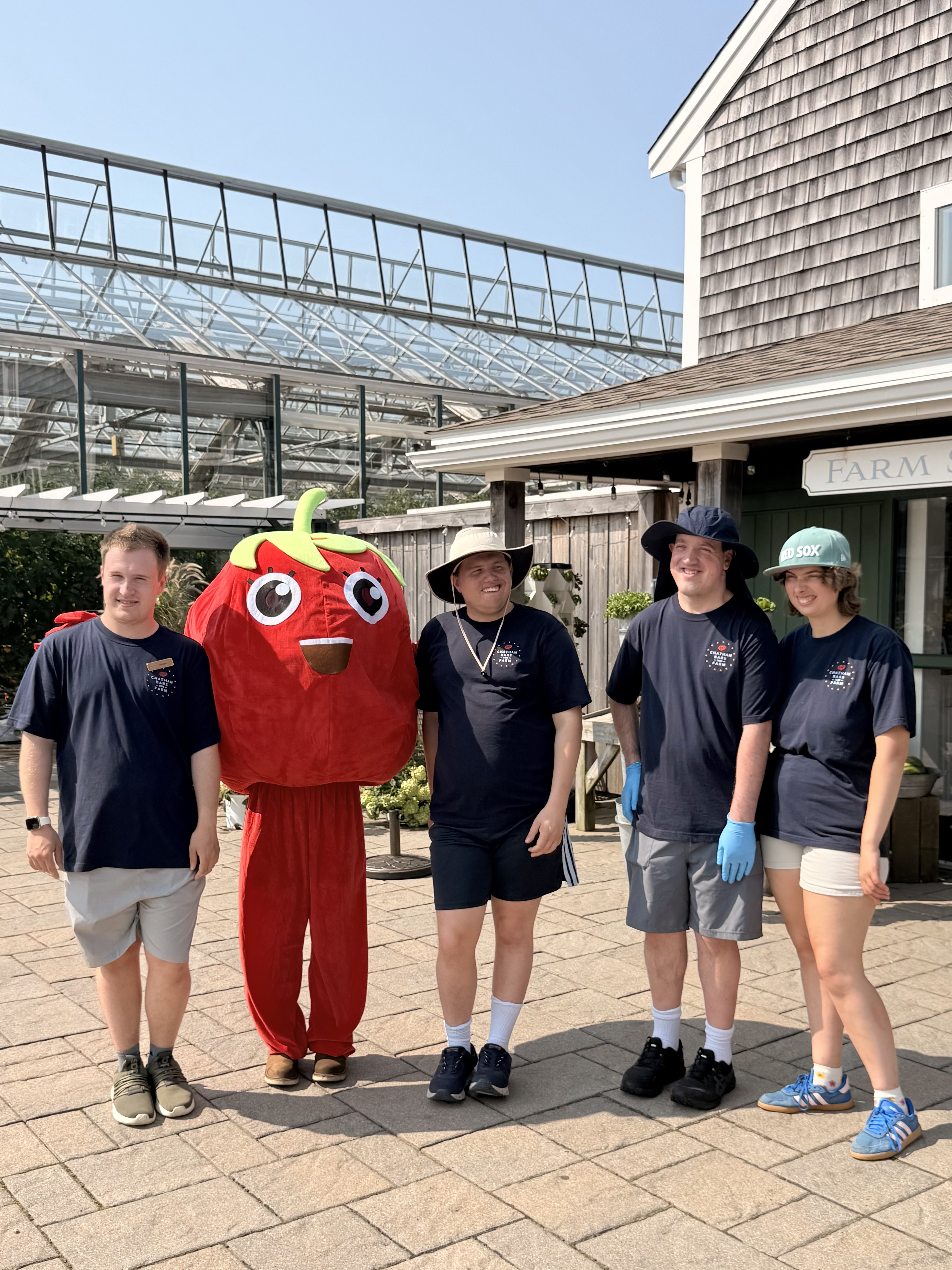 Five people stand together, with one in a tomato costume. They're outside a building labeled "FARM" under sunny skies.