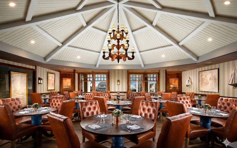 Luxurious restaurant interior with leather chairs, round tables set with napkins, a central chandelier, and nautical-themed decor.
