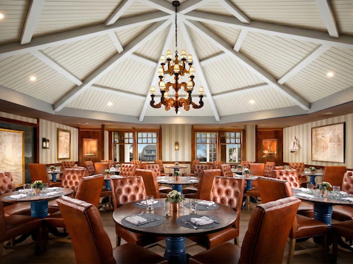 A restaurant dining area with round tables, brown leather chairs, a central chandelier, and decorative walls and ceiling.