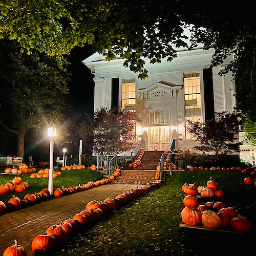 A well-lit building with a grand staircase, surrounded by numerous pumpkins arranged decoratively on the pathway and lawn.