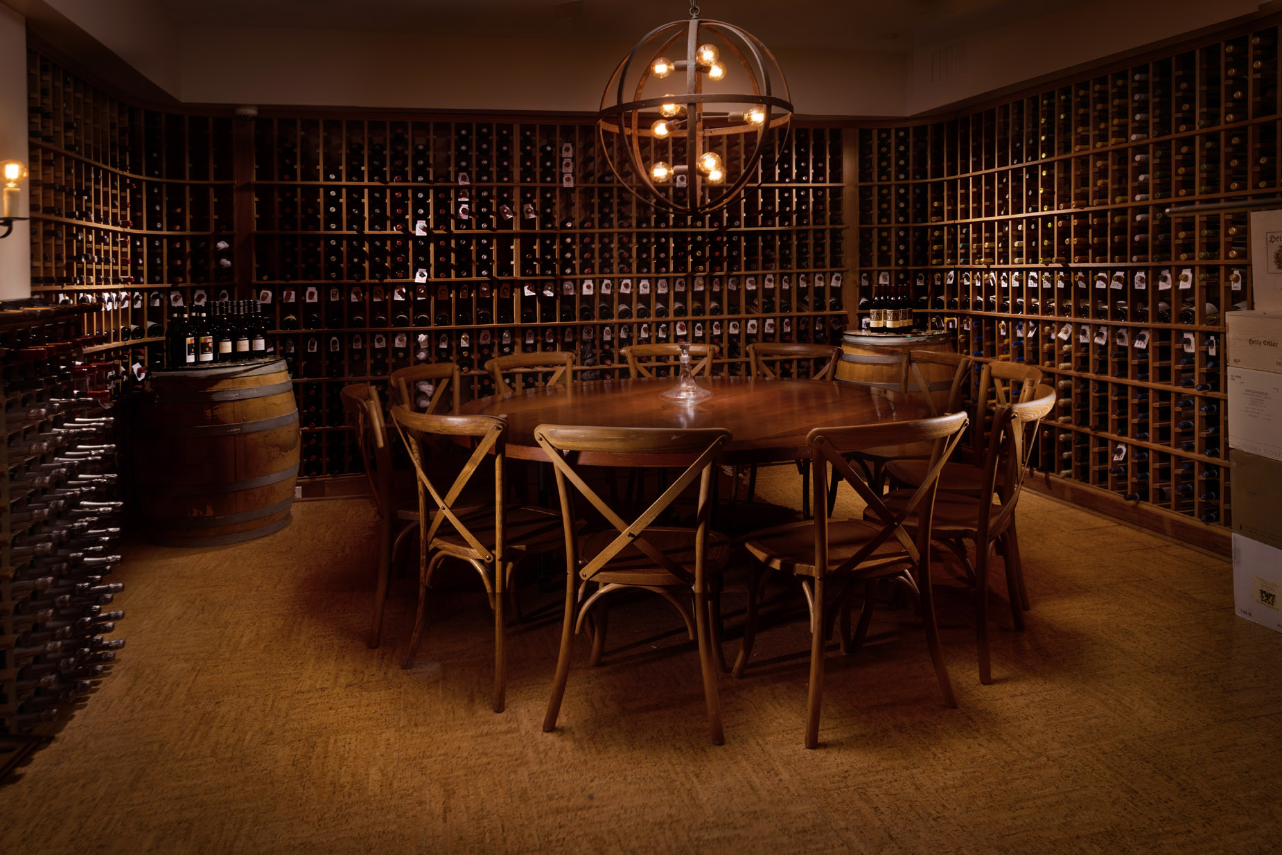 A wine cellar with wooden shelves full of wine bottles, a central wood table surrounded by chairs, and barrels used as stands for more bottles.