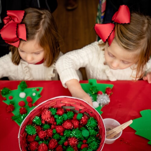 Children are crafting with holiday-themed decorations at a table, featuring foam trees and pom-poms.