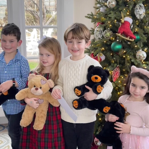 Children are standing in front of a Christmas tree holding stuffed animals, with festive decorations around.