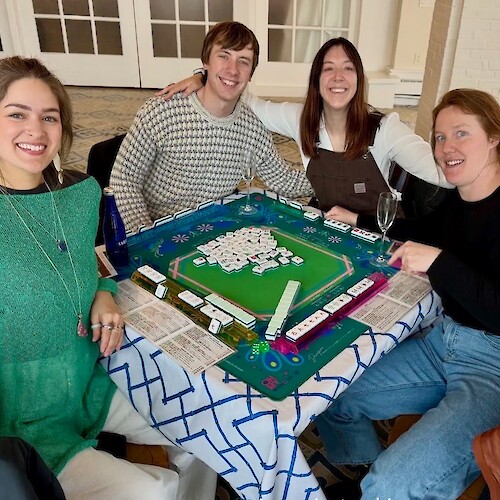 Four people are smiling around a table during a game of mahjong, with tiles and scorecards visible.