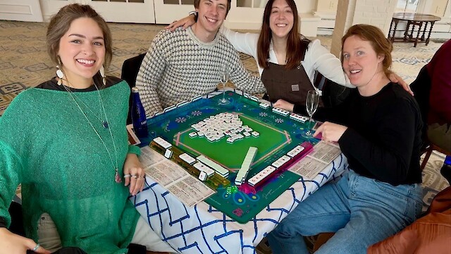 Four people are smiling around a table during a game of mahjong, with tiles and scorecards visible.