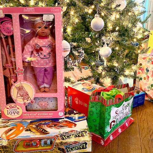 A Christmas tree surrounded by various wrapped gifts, including a doll stroller set, a toy dinosaur set, and festive bags.