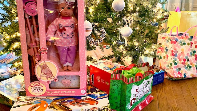 A Christmas tree surrounded by various wrapped gifts, including a doll stroller set, a toy dinosaur set, and festive bags.