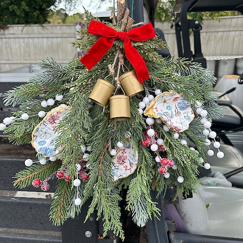 A festive arrangement with pine branches, a red ribbon, gold bells, and ornaments hangs on a vehicle.
