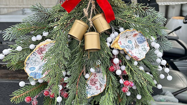 A festive arrangement with pine branches, a red ribbon, gold bells, and ornaments hangs on a vehicle.