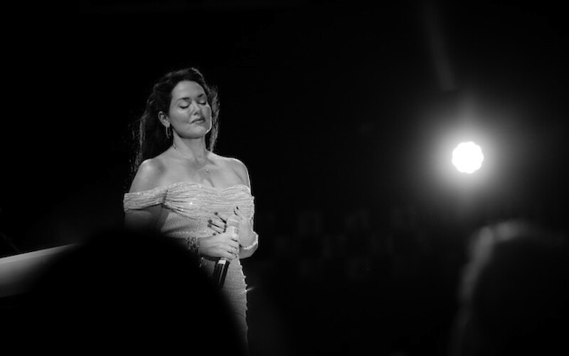 A person stands on stage in a spotlight, holding a microphone, with eyes closed, in a black and white setting.