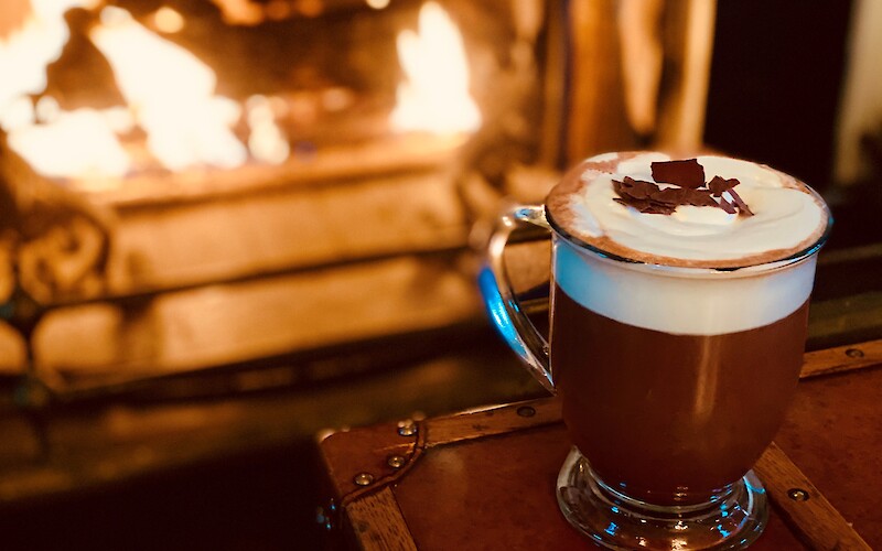 A cozy scene with a glass mug of hot chocolate topped with whipped cream and chocolate shavings, set by a warm, glowing fireplace.