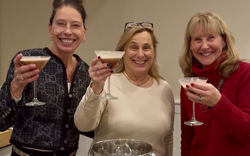 Three people smiling and holding drinks stand around a table with barware and an ice bucket.