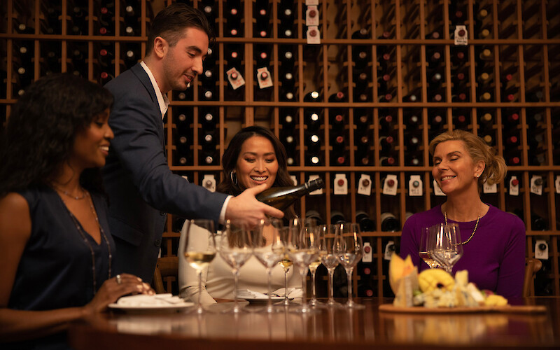 A group at a wine tasting; a man pours wine as women enjoy the setting, surrounded by wine racks.