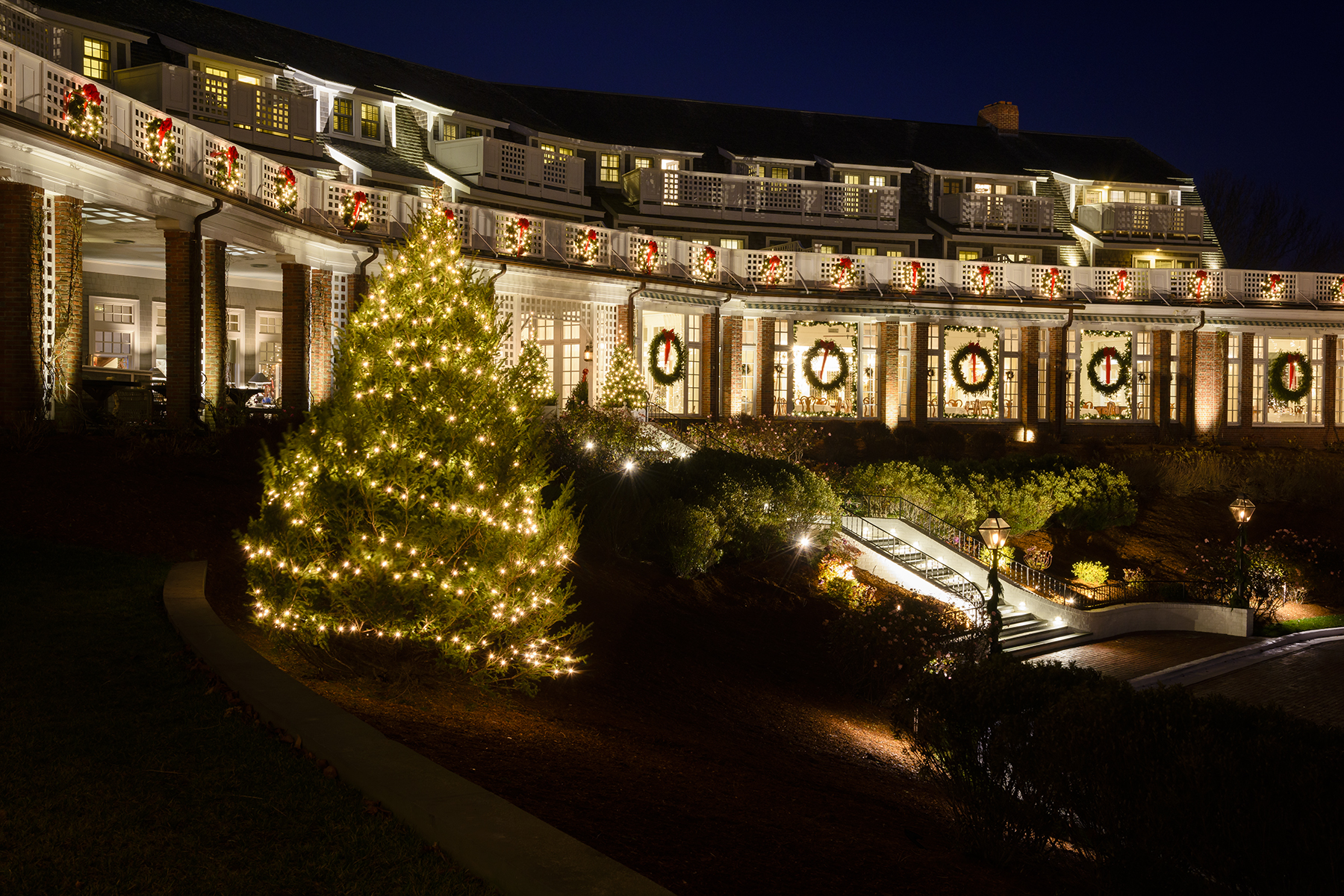 A building decorated with Christmas wreaths and bows, surrounded by snow, bushes, and a bench in the foreground of the image.