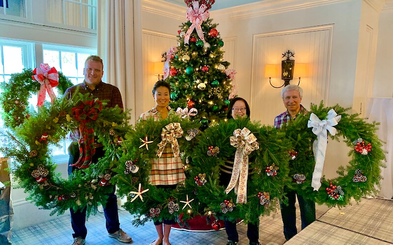 Four people stand holding large Christmas wreaths in front of a decorated Christmas tree indoors.
