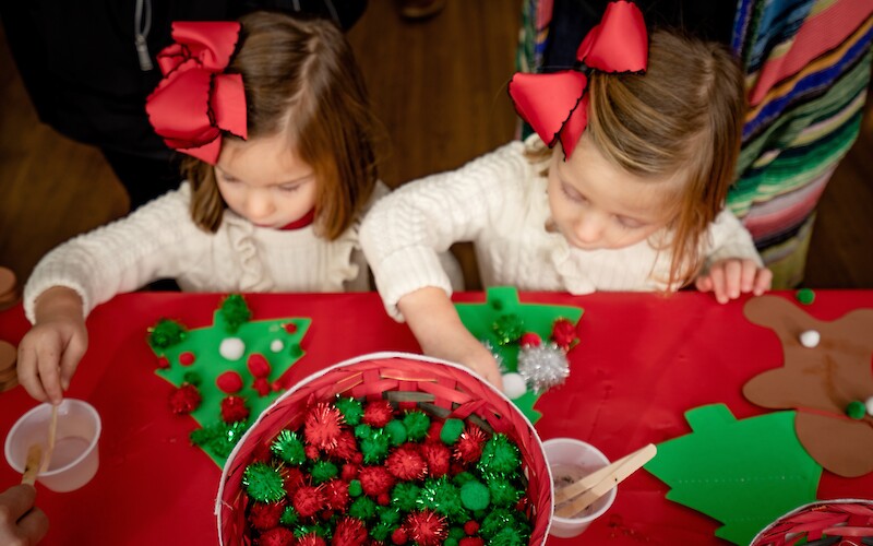 Two children with red bows are crafting at a table decorated with red and green materials and a basket of pom-poms.