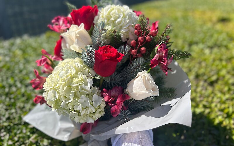 A bouquet with red and white flowers, greenery, and berries, held against a blurred green background in sunlight.