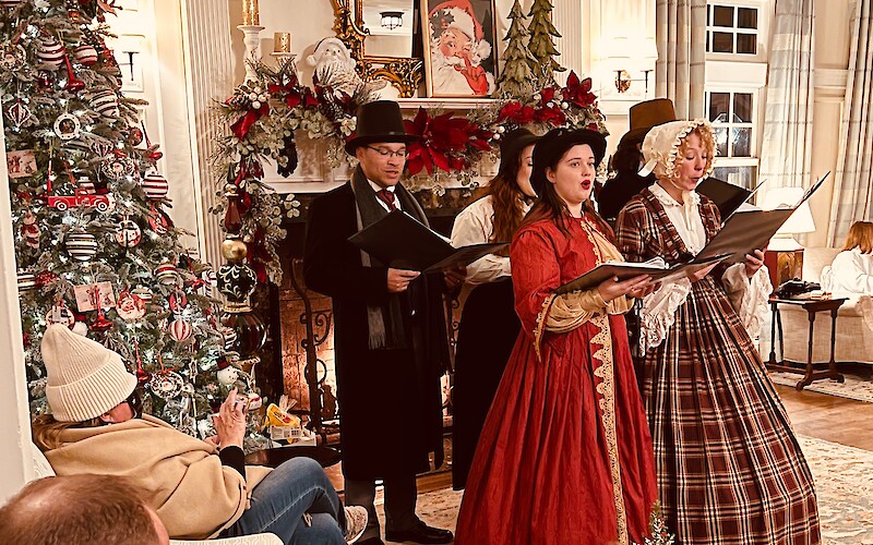 A group in period attire sings by a festive fireplace, surrounded by onlookers and a decorated Christmas tree.