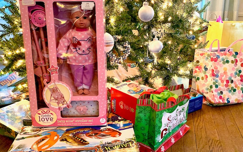 Christmas gifts under a tree, featuring a doll set, toy trucks, and colorful gift bags with festive decorations.