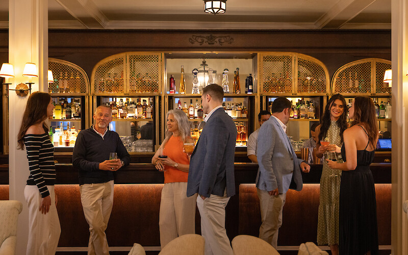 A group of people enjoying drinks at a stylish bar, engaged in lively conversation, with bottles displayed on shelves behind them.