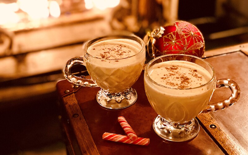 Two mugs of creamy beverage topped with spice, a red ornament, candy canes, and a fireplace in the background.