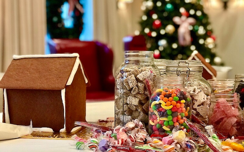 A gingerbread house setup with jars of colorful candies and sweets, holiday decorations, and a Christmas tree in the background.